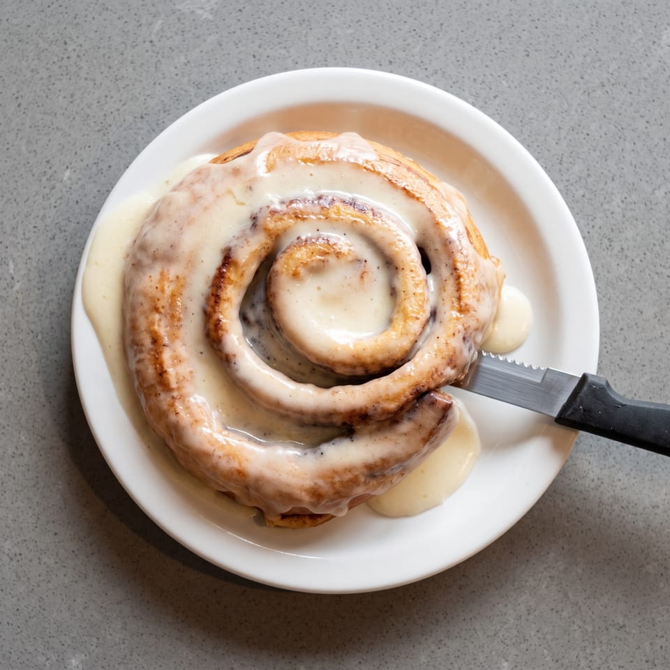 A large, frosted cinnamon roll on a white plate with a knife cutting into it.