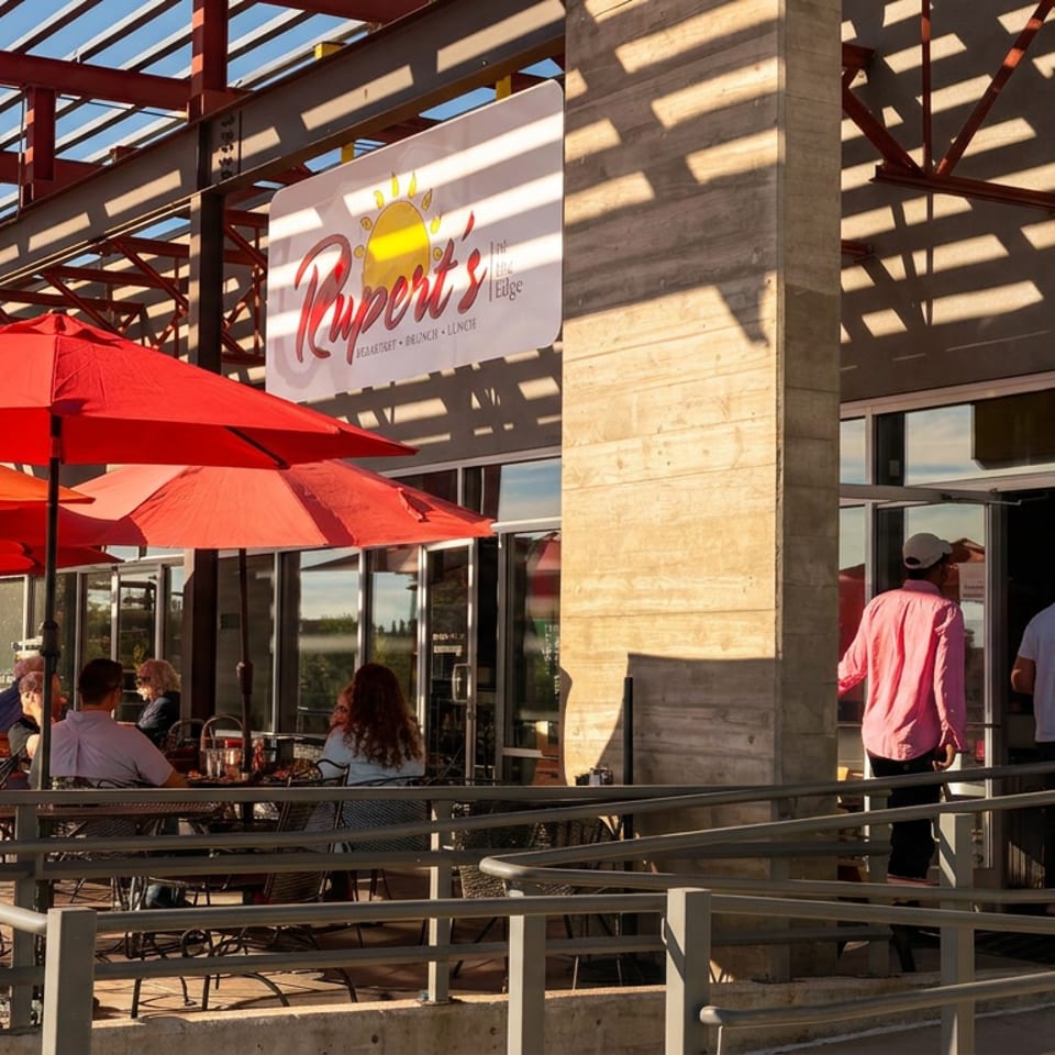 People dining on Rupert's at The Edge sunny outdoor patio with red umbrellas.