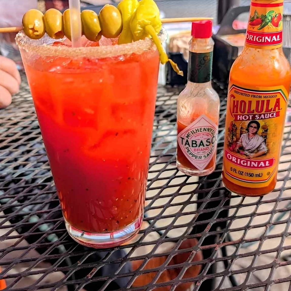 A Bloody Mary cocktail garnished with olives and peppers, alongside Tabasco and Cholula hot sauces.