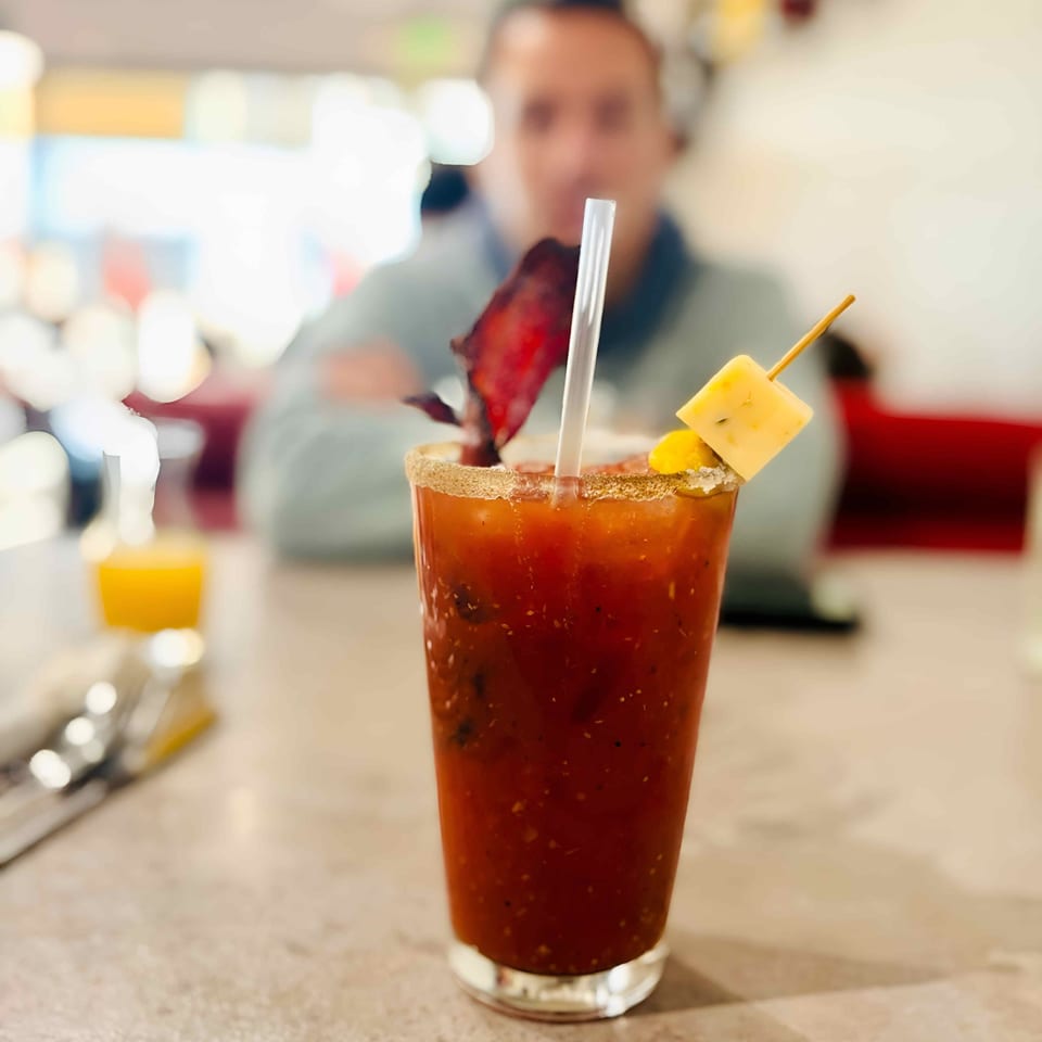A Bloody Mary cocktail with bacon, cheese, and a straw, on a table with a person in the background.