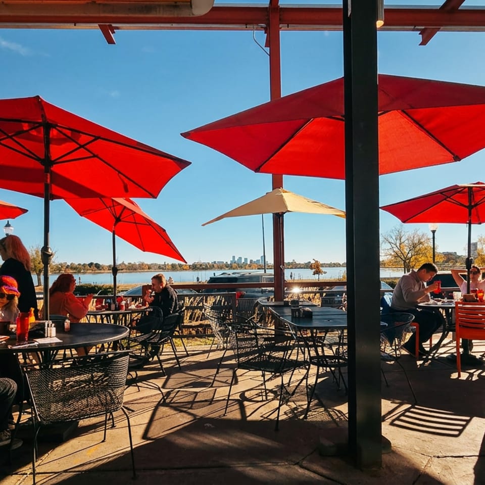 People dining outdoors on a sunny patio with red umbrellas overlooking a lake and city skyline.