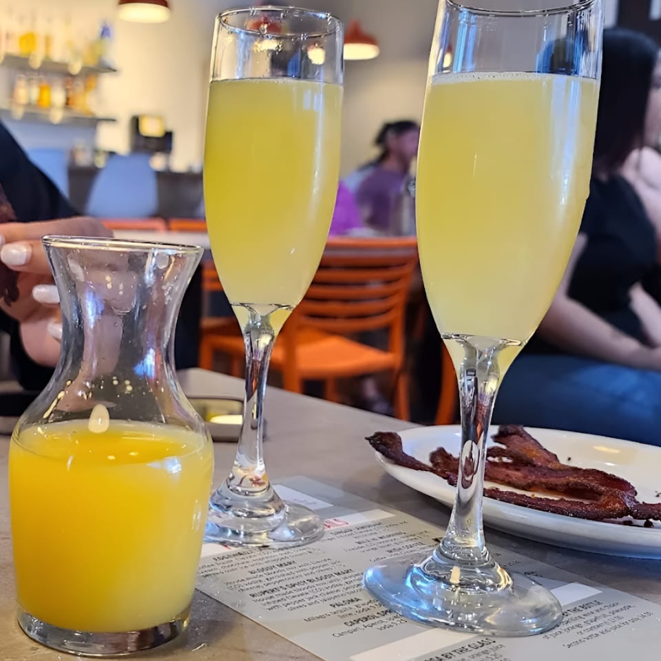 Two mimosa flutes and a carafe of orange juice on a table with bacon and a menu.