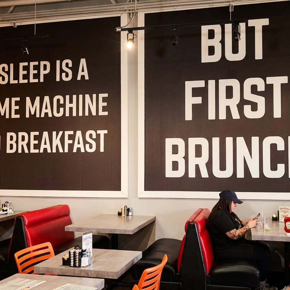 Restaurant interior with red booths, orange chairs, and two women under large "Breakfast" and "Brunch" signs.